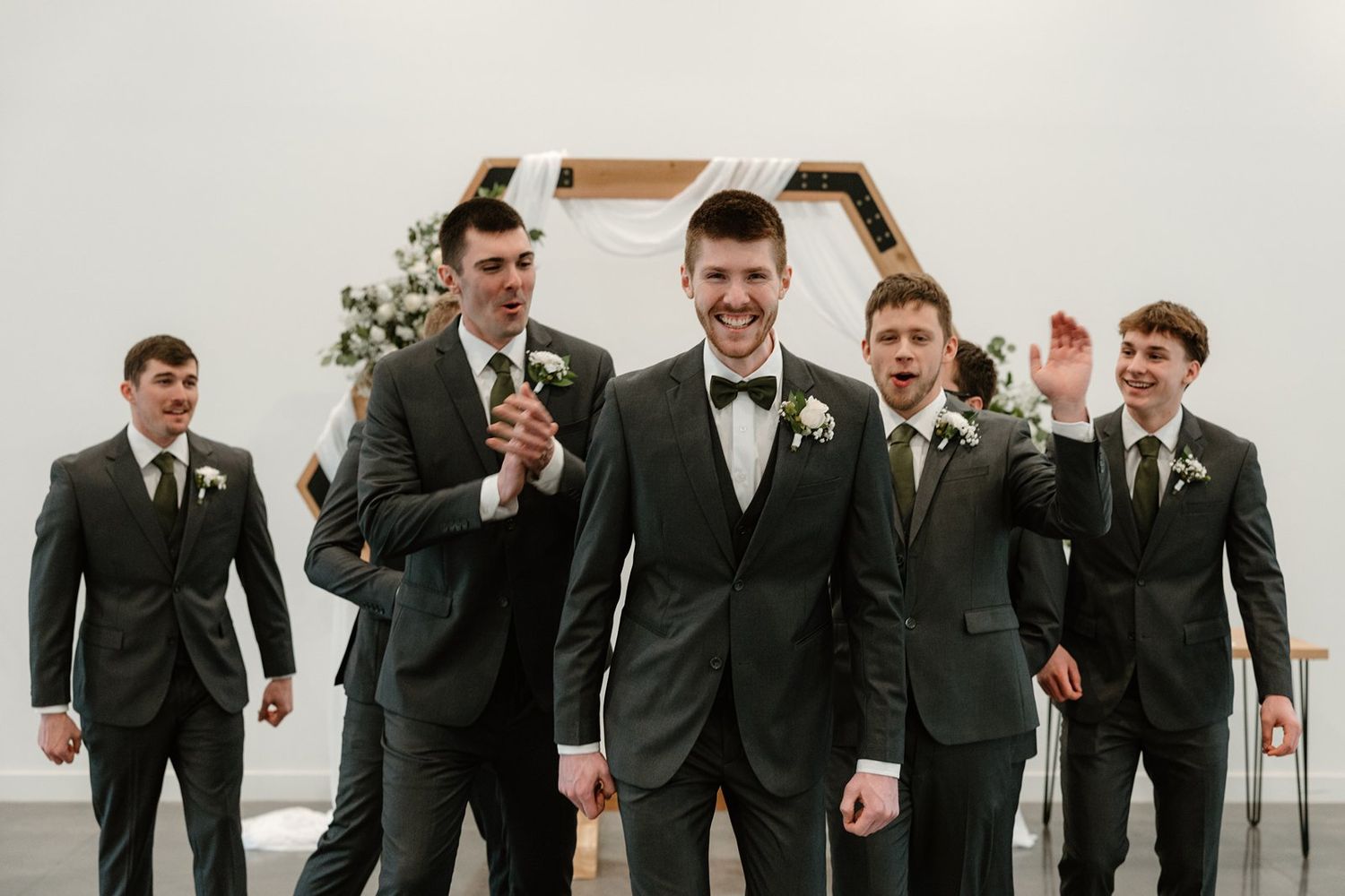 Groomsmen in matching black suits and bowties smile and celebrate at a wedding venue.