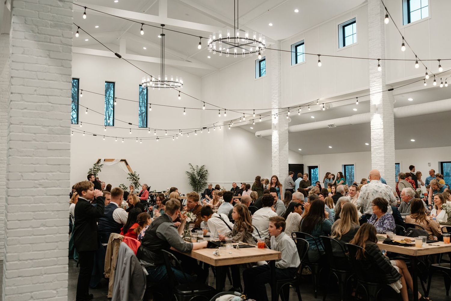 A large group of people gathered in a modern white-walled event space with track lighting and communal tables.