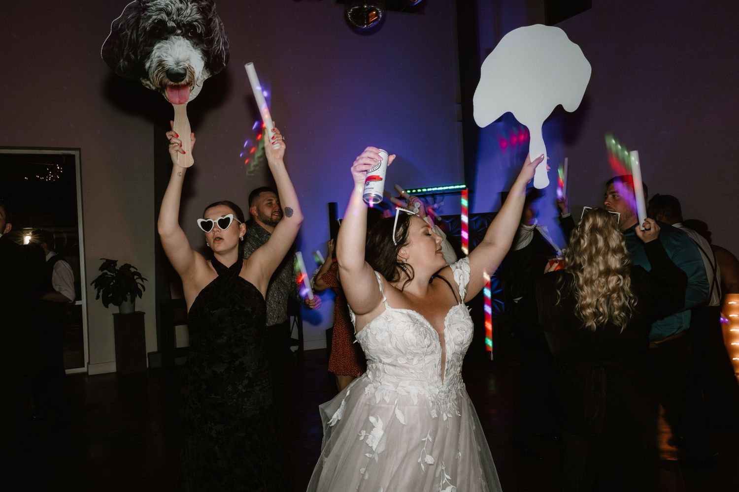People dancing and celebrating at a wedding reception with glow sticks and festive energy on a dark dance floor.