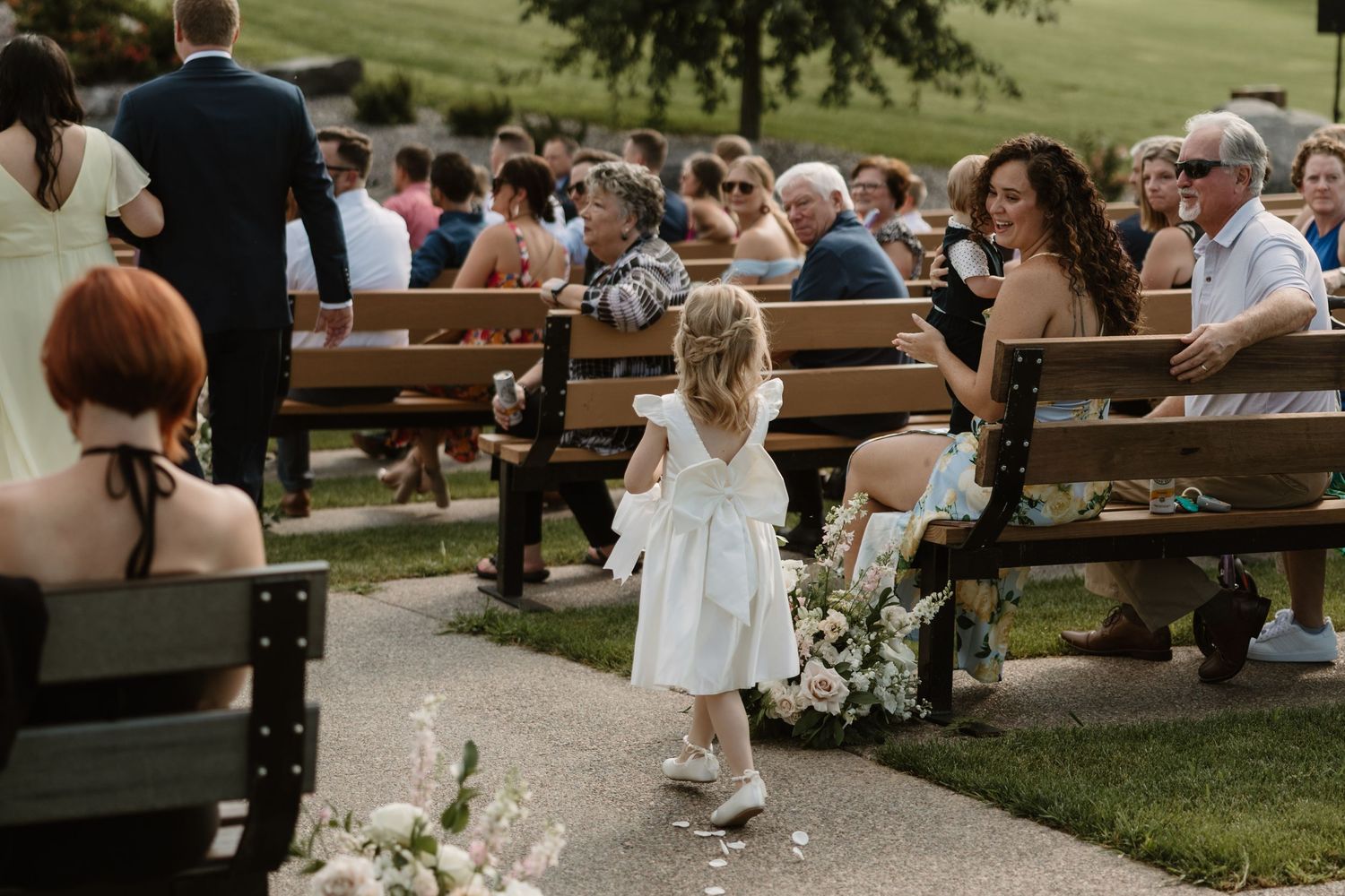Wedding guests gather outdoors on wooden benches during a summer celebration in a garden setting.