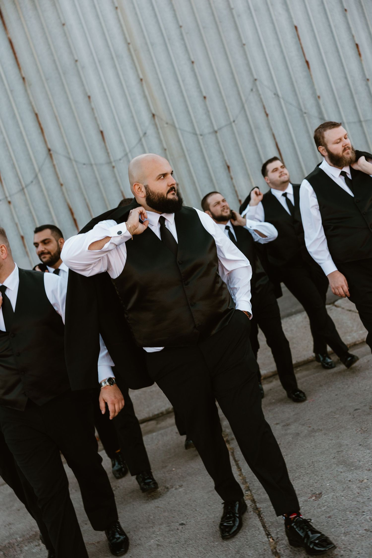 Groomsmen in matching black and white formal attire pose together against an industrial wall background.