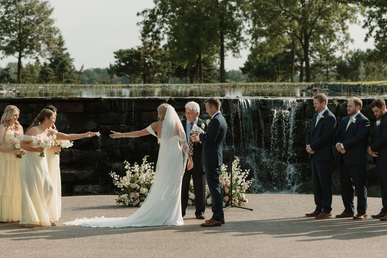 A wedding ceremony takes place outdoors with a scenic waterfall backdrop and wedding party lined up on a stone patio.