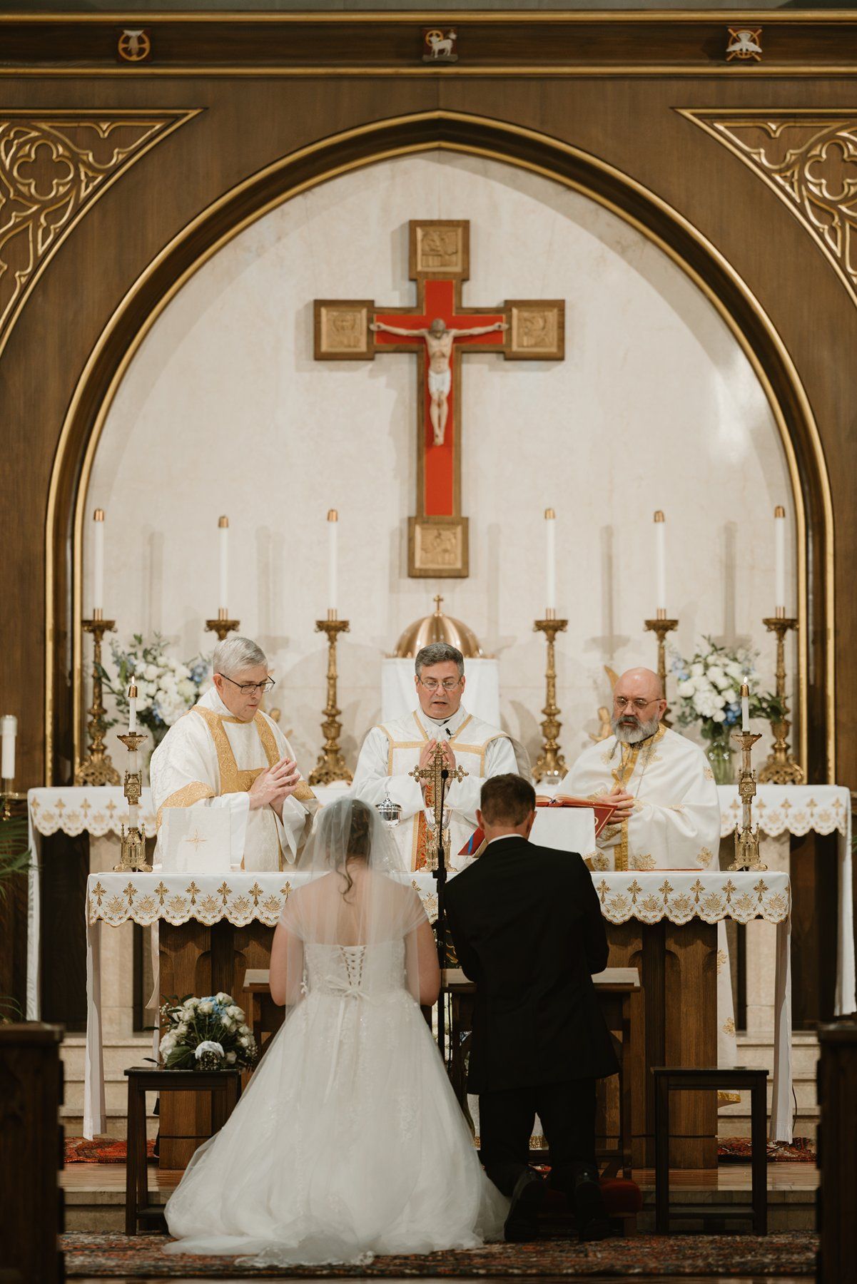 A traditional Catholic wedding ceremony takes place at an altar with a crucifix, candles and floral decorations in a church.