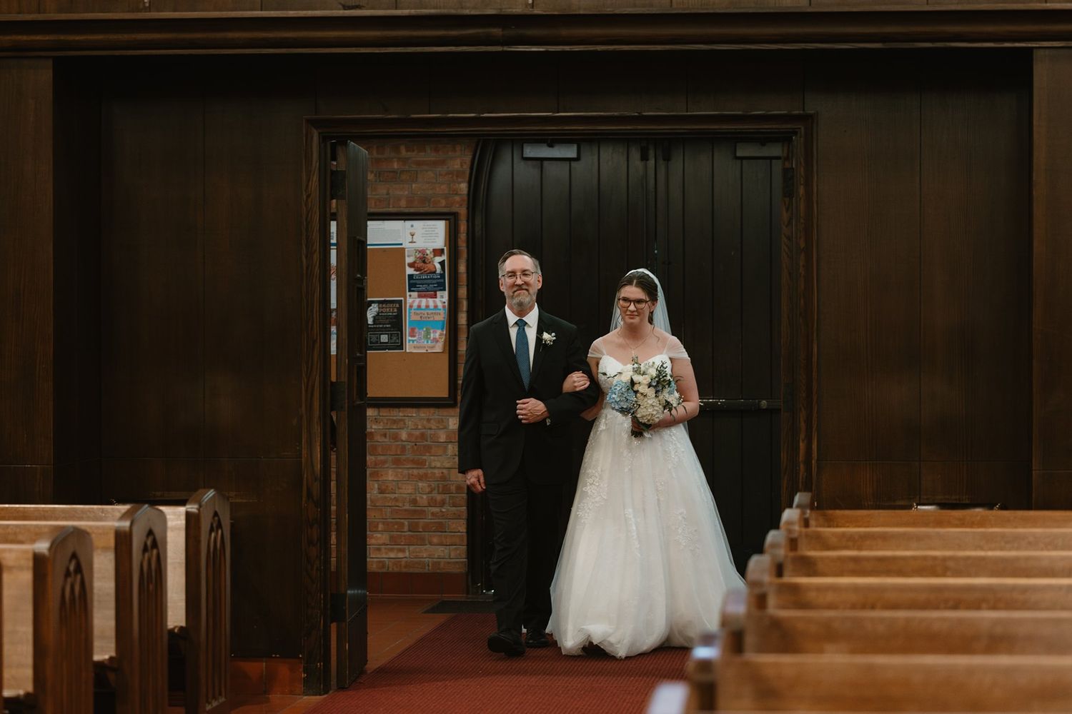 A bride and her escort pause at the entrance of a traditional wooden church with empty pews in view.