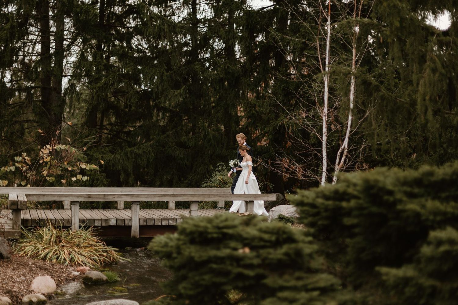 A bride in a white gown walks across a wooden bridge surrounded by evergreen trees.