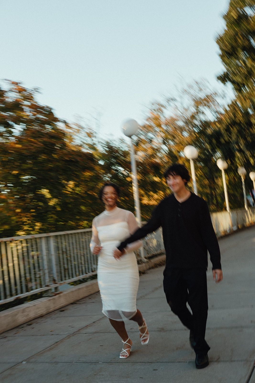 A joyful couple walks hand-in-hand on a waterfront path, woman in white dress, man in black outfit, with motion blur effect.