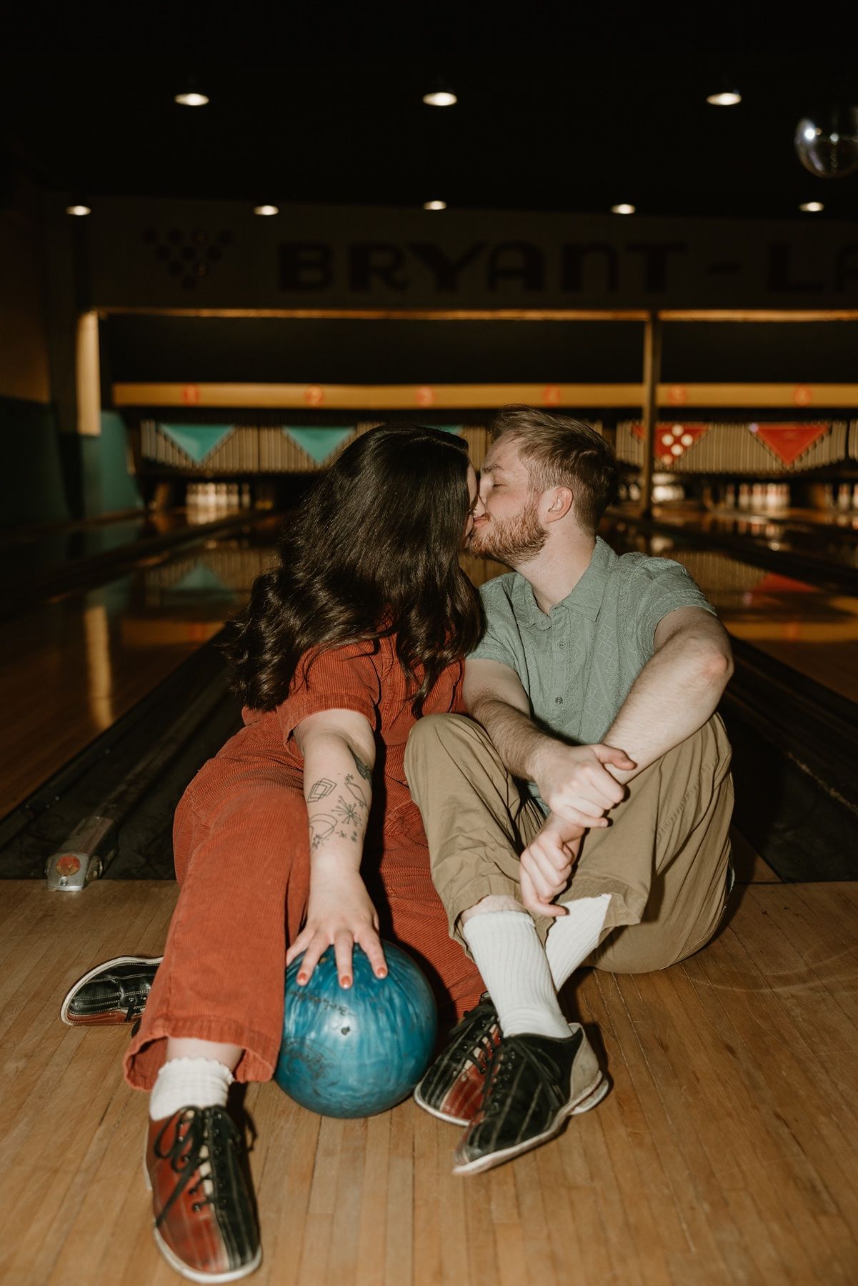 A couple shares a romantic moment while sitting on a bowling alley lane with a teal bowling ball between them.