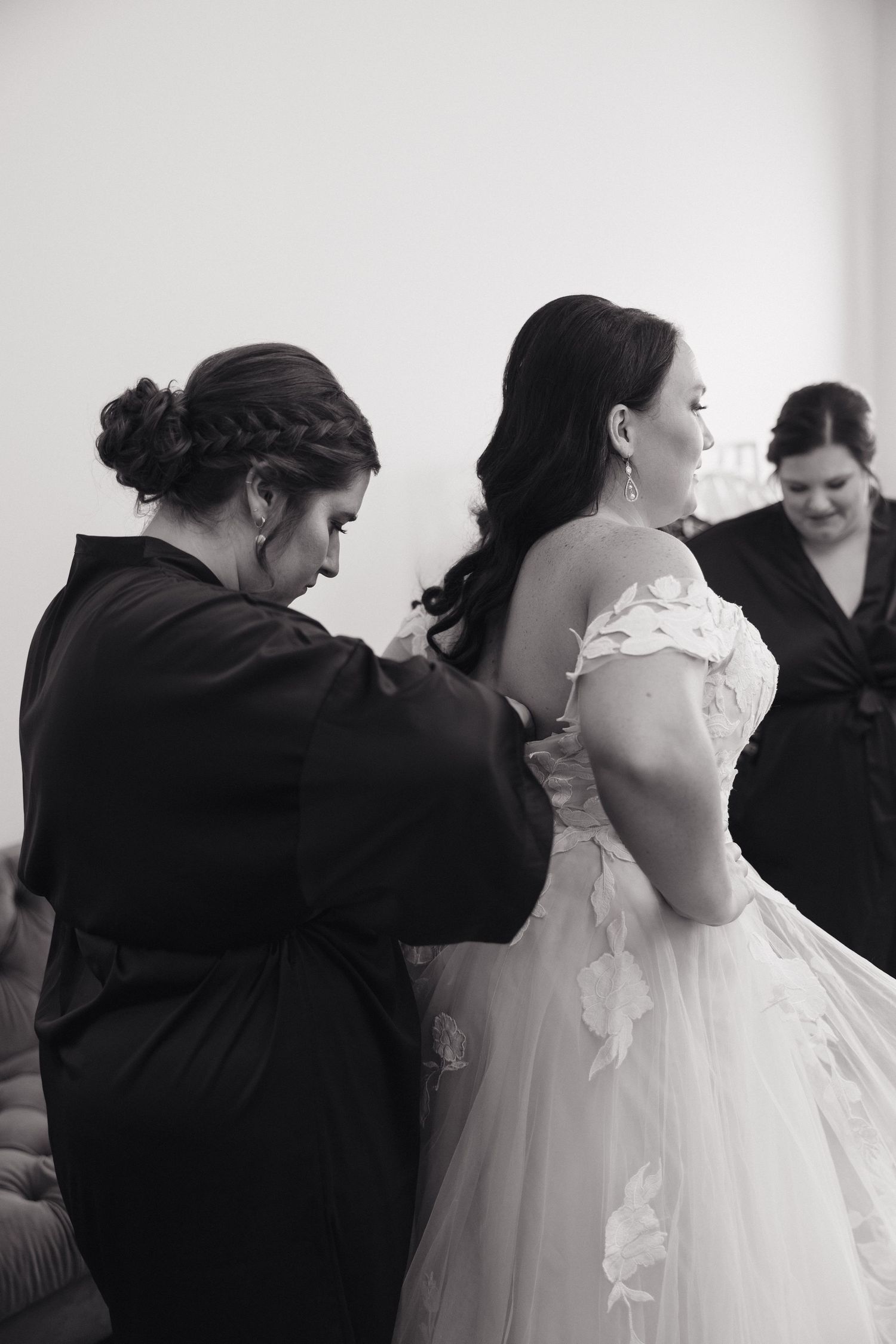 A bride in a white wedding gown gets help with final dress adjustments during wedding preparations in black and white.