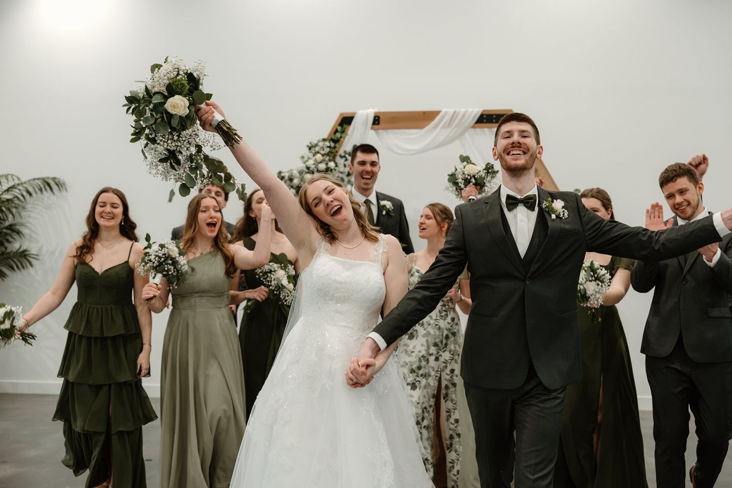 Wedding party celebrates joyfully outdoors in olive green and black formal attire.