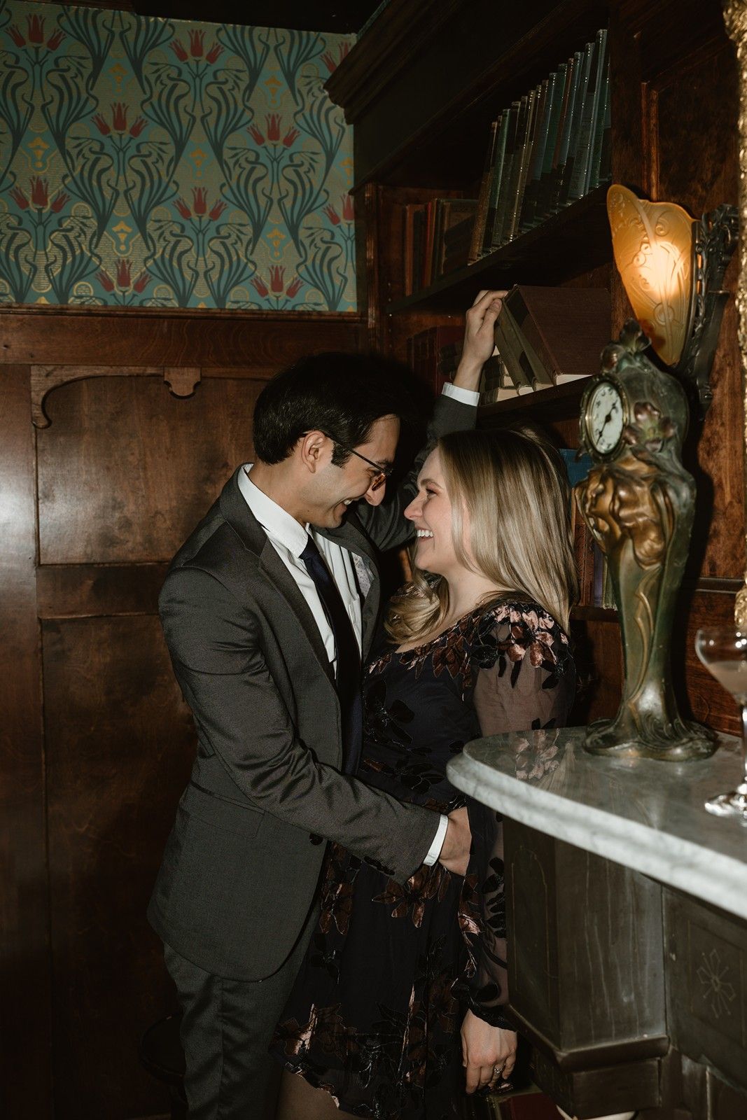 A romantic couple in formal attire share an intimate moment near an ornate lamp against vintage wallpaper decor.