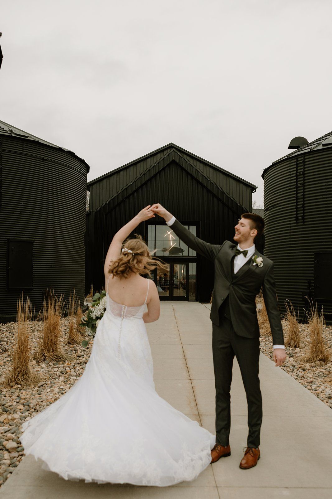 A couple in wedding attire dances and twirls together on a concrete path near industrial storage silos.