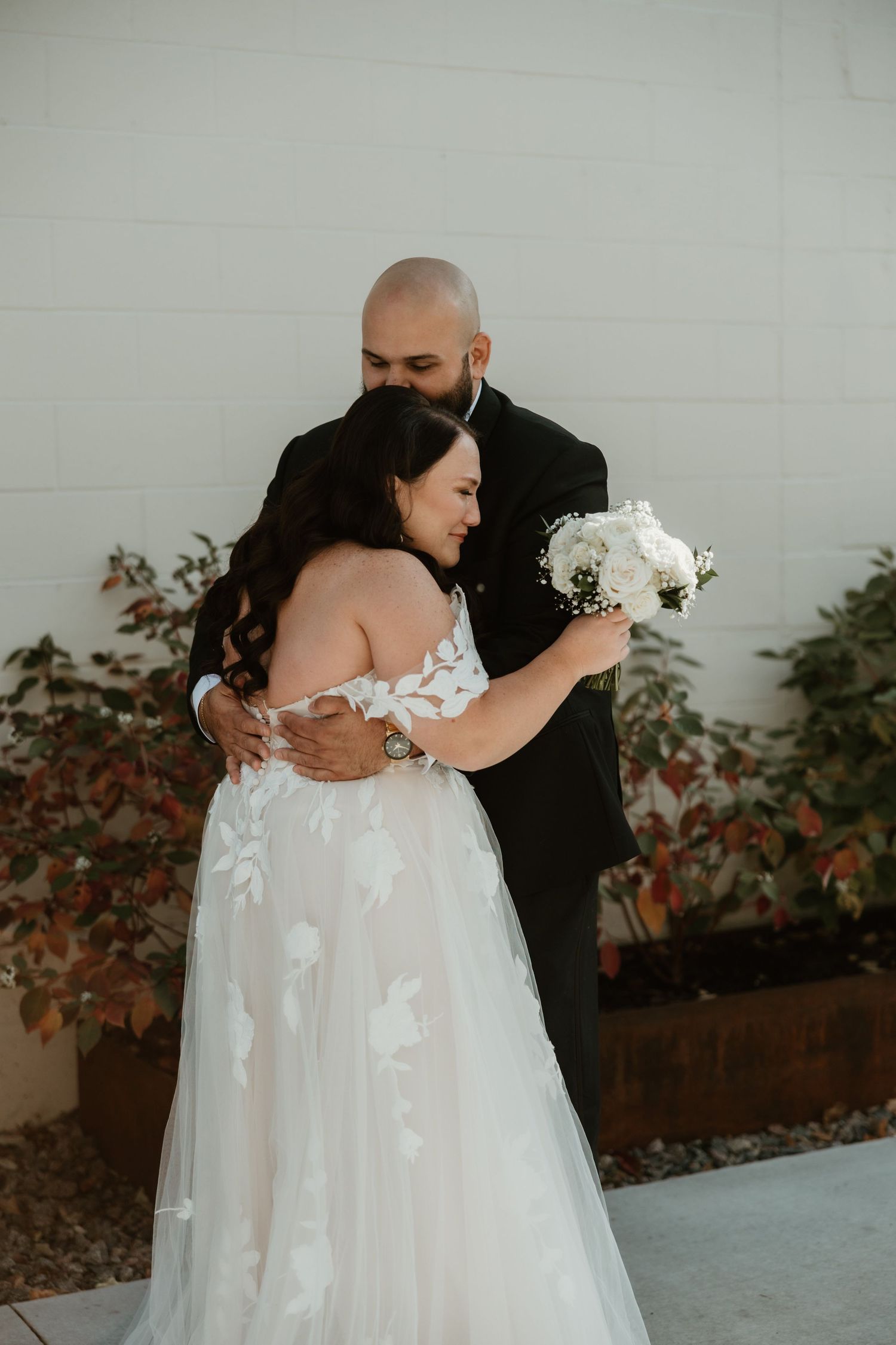 A couple embraces at their wedding, the bride wearing a white off-shoulder gown holding a bouquet of white flowers.