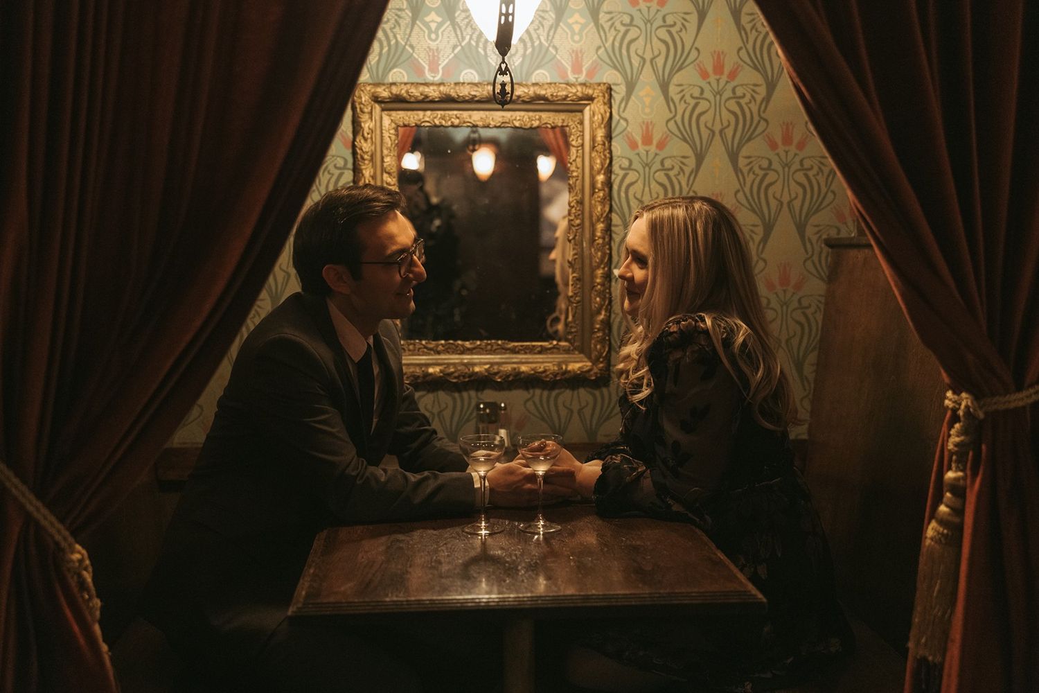 Two people enjoy drinks at a table in a dimly lit restaurant with ornate wallpaper and dark curtains.