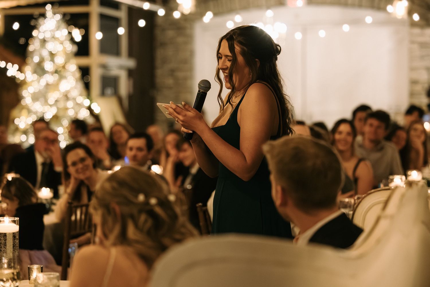 Woman in dark green dress giving a speech at an elegant wedding reception with warm string lights.
