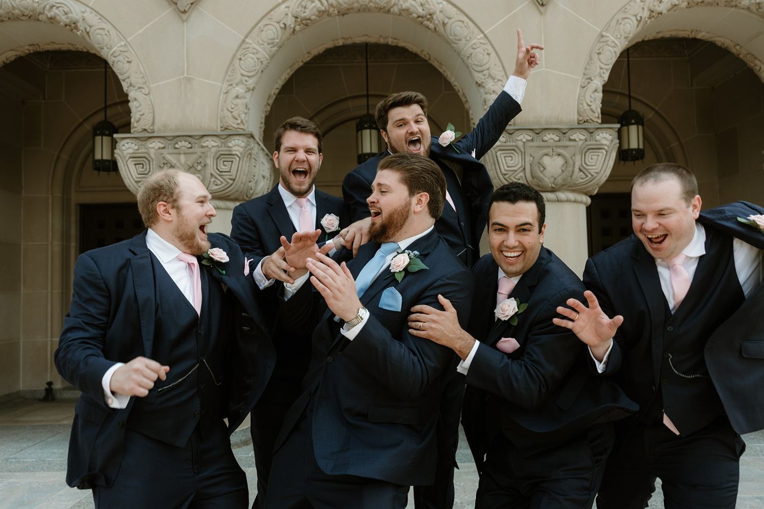 Groomsmen in black suits share a playful moment hugging and laughing together in an elegant archway setting.