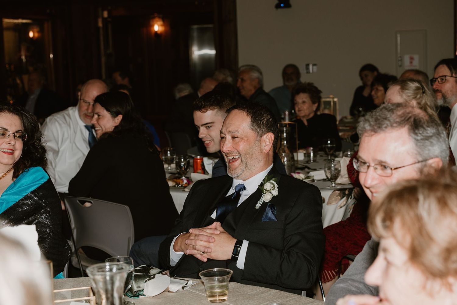 Guests laugh and smile while seated at tables during an indoor wedding reception.