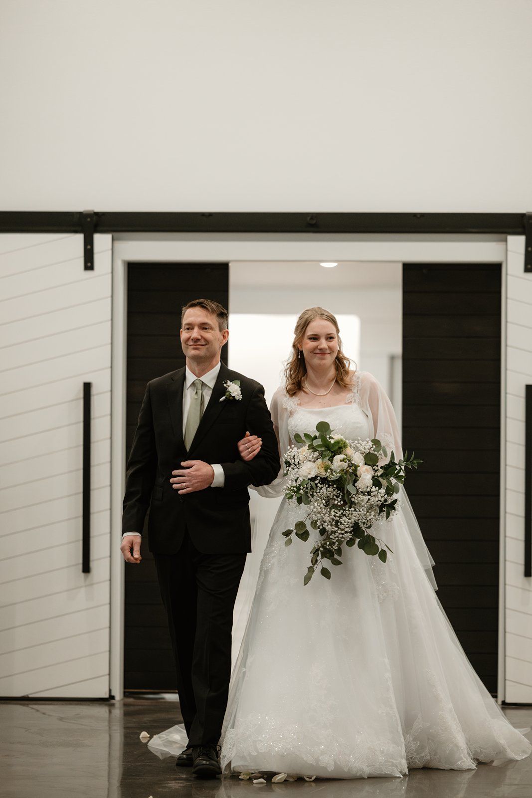A wedding couple in formal attire stands in a modern white hallway with black doors.