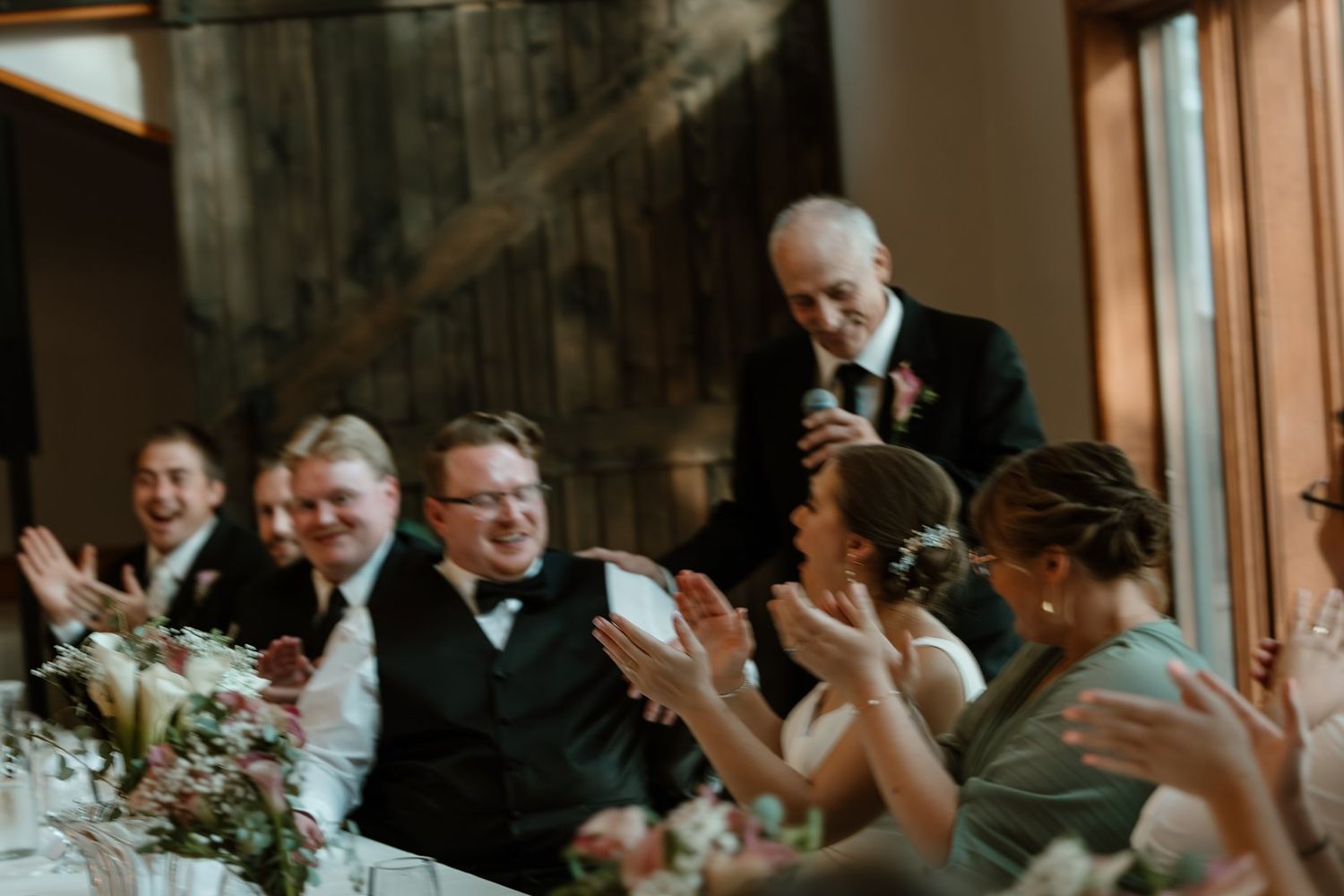 Wedding guests raise their glasses in celebration during an indoor reception at a venue with large windows.