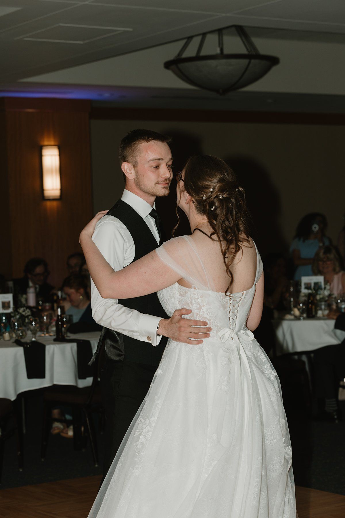 A couple shares their first dance at their wedding reception in a dimly lit ballroom with white decor.
