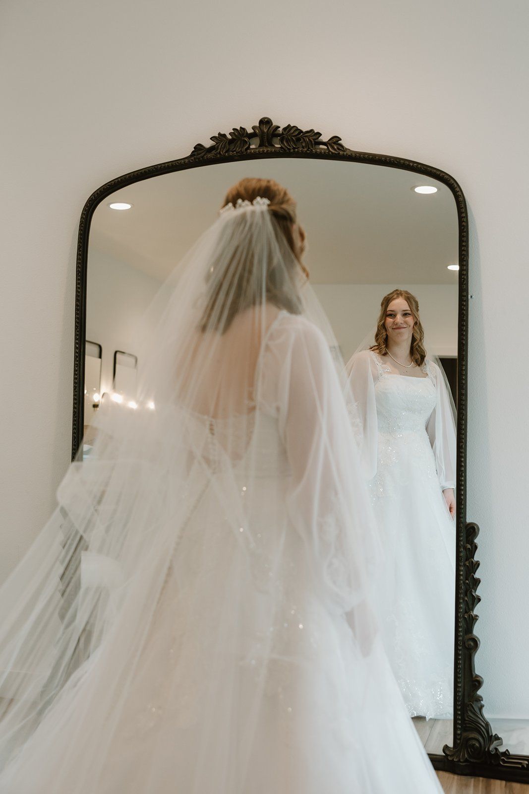 A bride in a flowing white wedding gown and long veil poses in front of a mirror in a bridal boutique.
