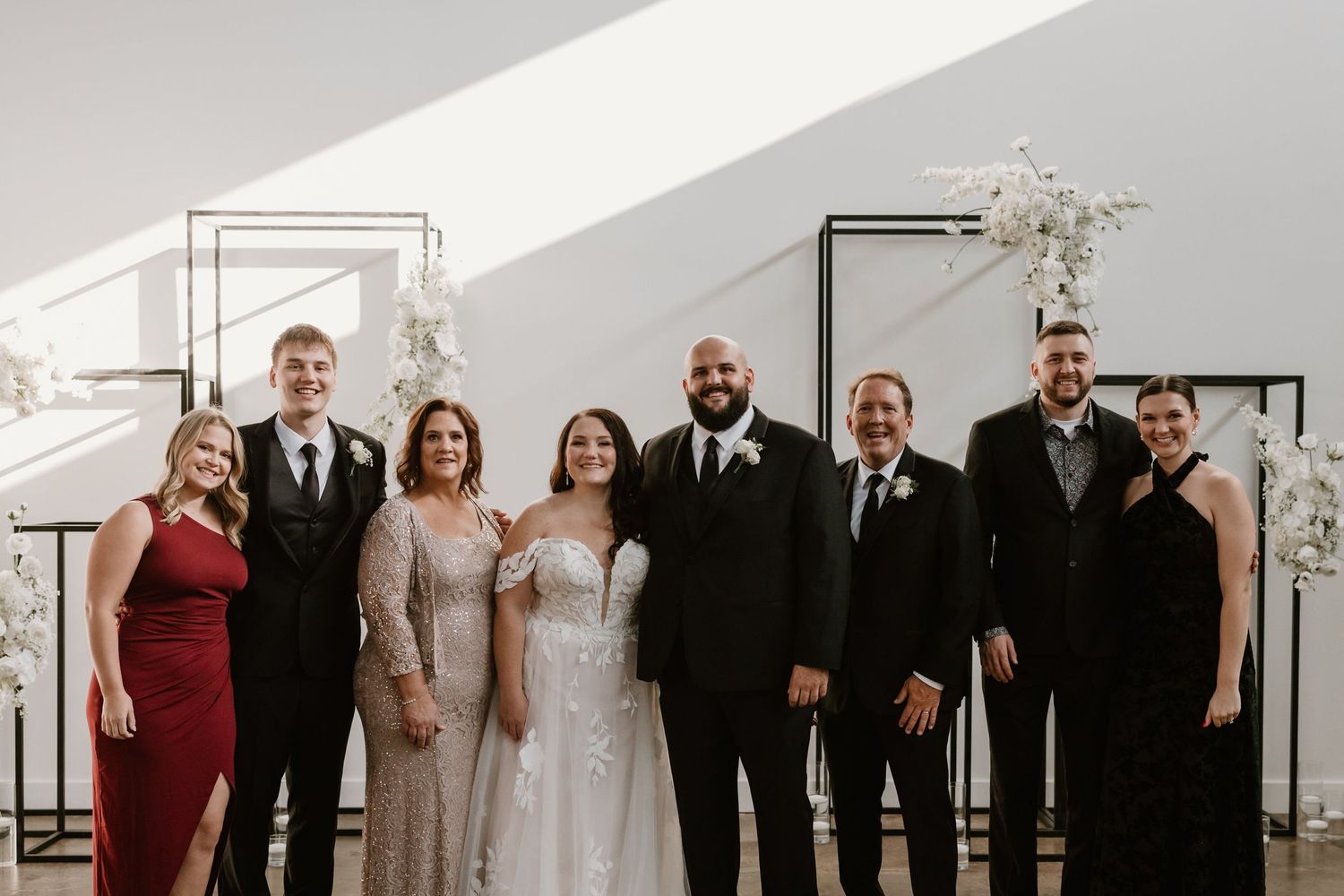A wedding party in formal attire poses together against a white wall with floral arrangements.