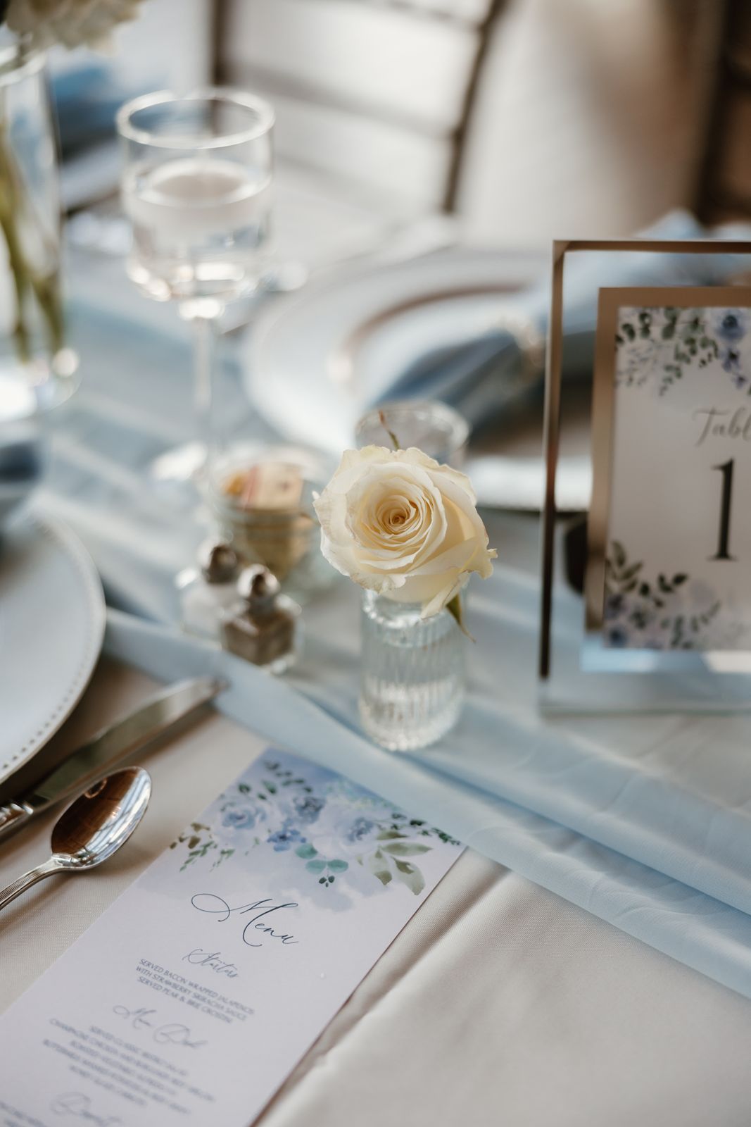 Close-up of wedding table decor with white rose, menu card and blue accent pieces.