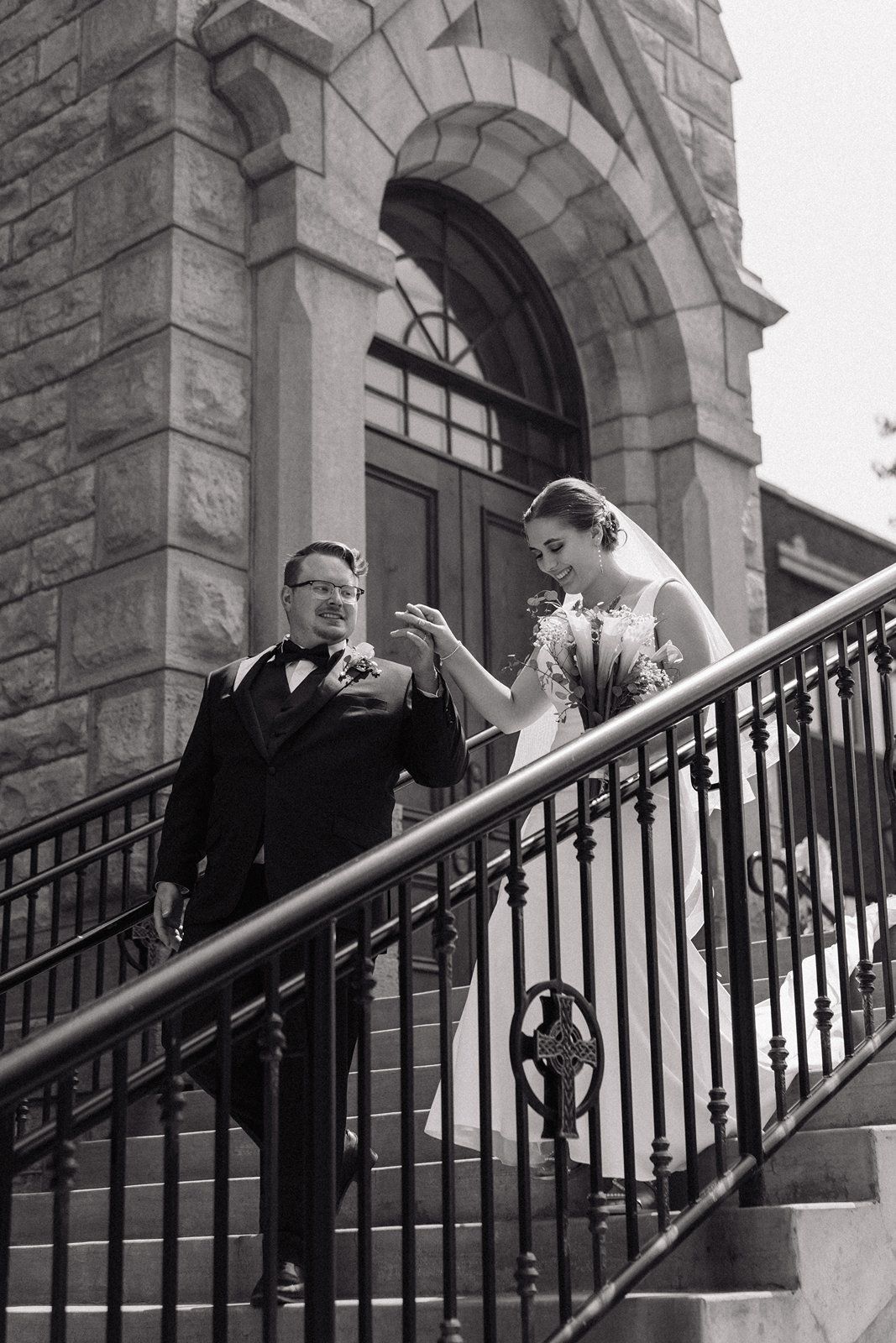 A couple stands on stone church steps with an arched doorway, sharing a joyful moment in black and white.