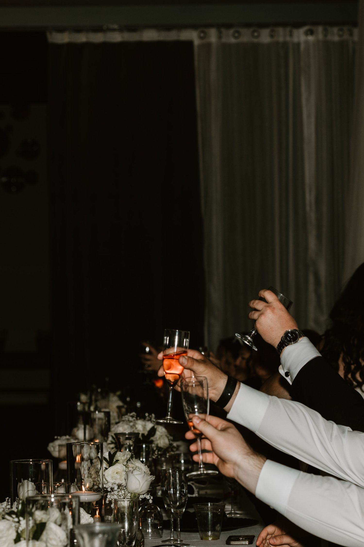 Hands in formal attire raise champagne glasses for a celebratory toast at an elegant dark dinner table.