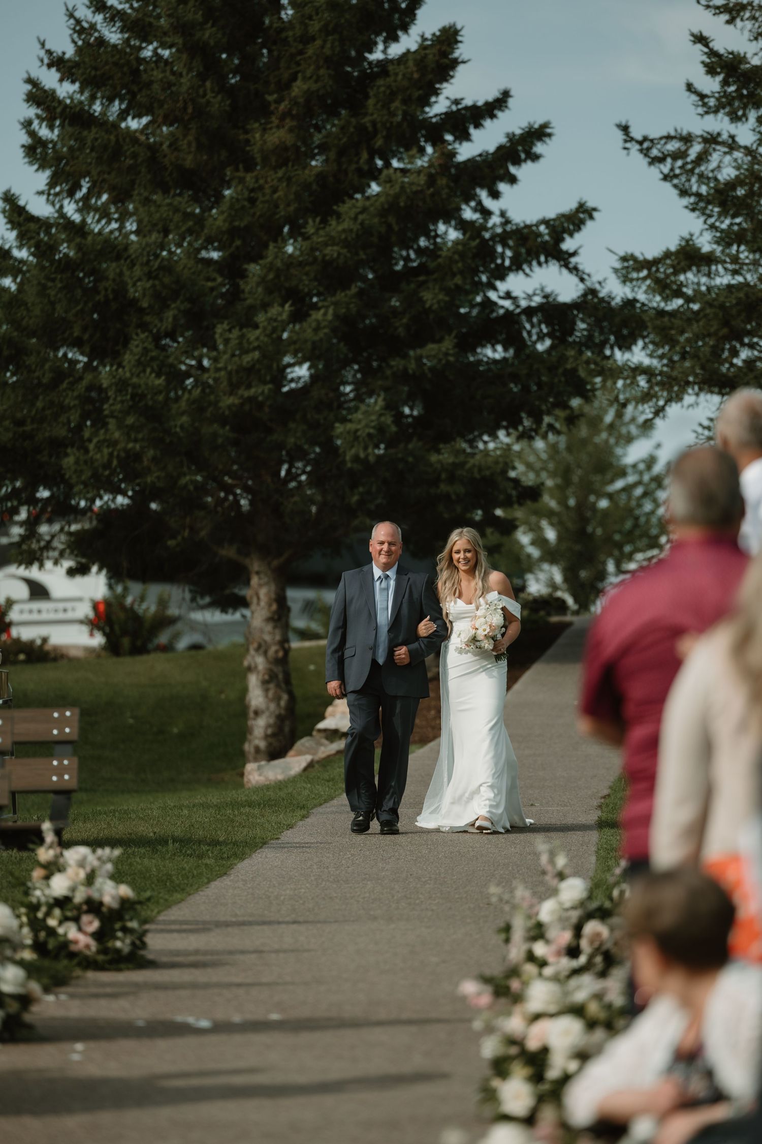 A wedding ceremony processional takes place on a path lined with evergreen trees and flowers on a sunny day.