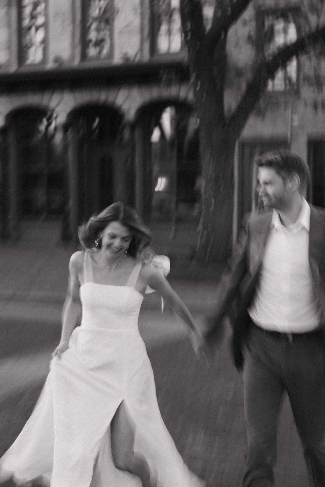 Black and white photo of a joyful bride and groom running together outdoors near historic arched building.