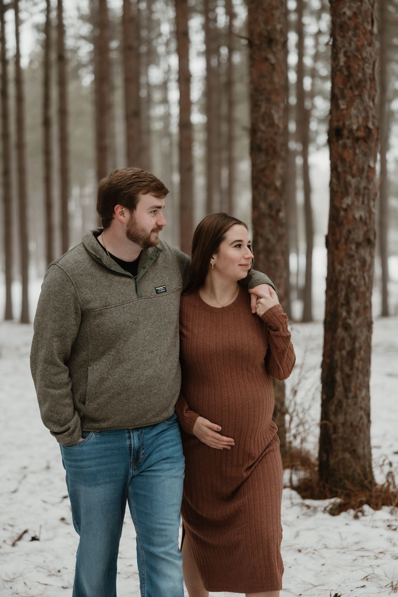 Couple posing in snowy pine forest, woman in brown ribbed maternity dress, man in gray pullover and jeans.