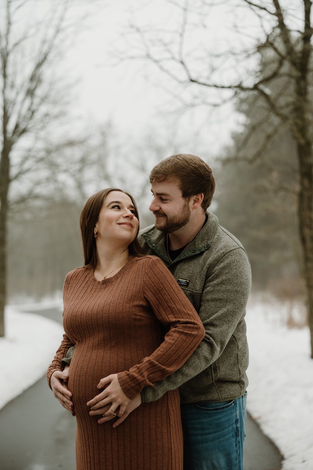 Couple embracing in snowy winter park, pregnant woman in brown dress smiling up at partner holding her belly.