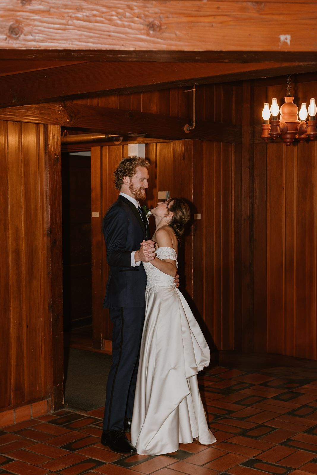 Couple shares their first dance in a wood-paneled room with warm lighting.