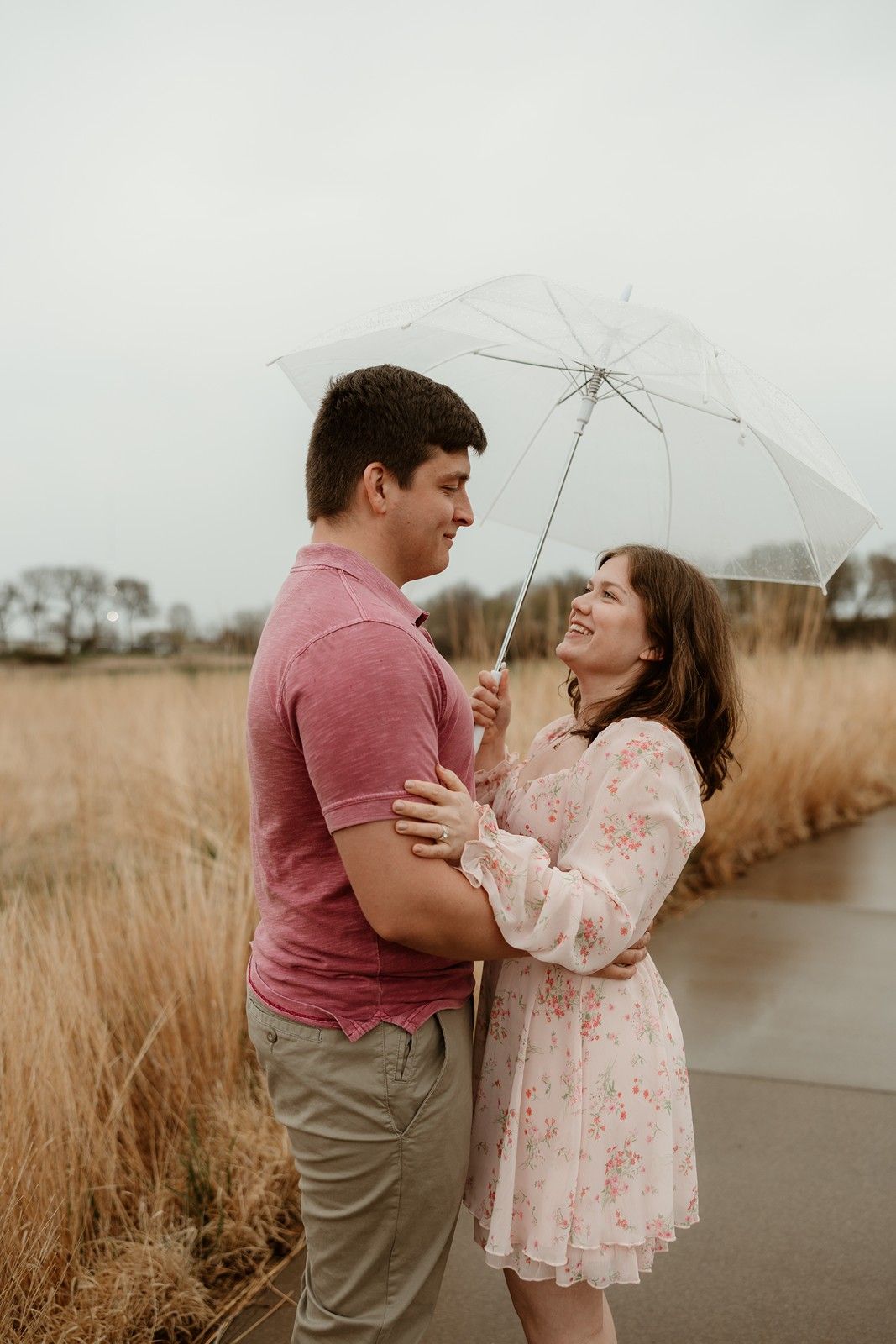 A couple shares a romantic moment under a clear umbrella on an overcast day by tall grass.