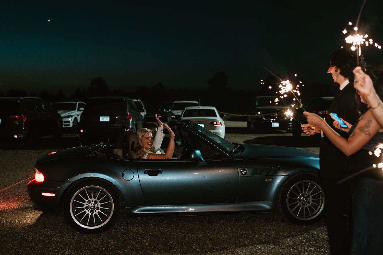 Newlyweds depart in a blue sports car while guests hold sparklers in the evening.