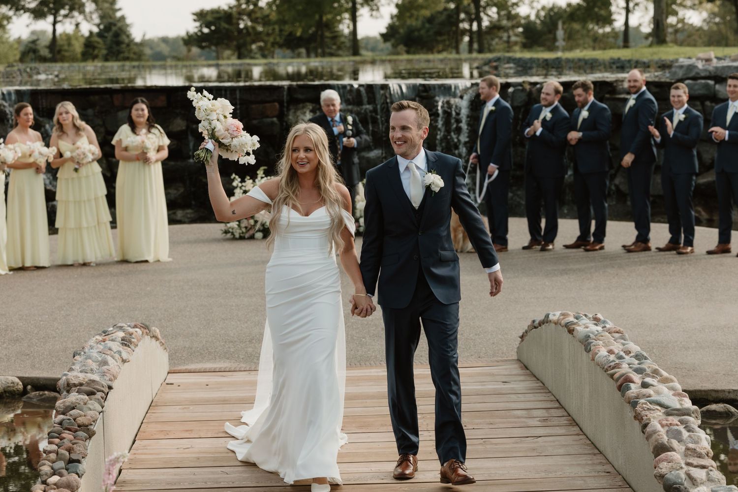 A couple walks hand in hand on a stone path at their outdoor wedding ceremony while bridal party members look on.