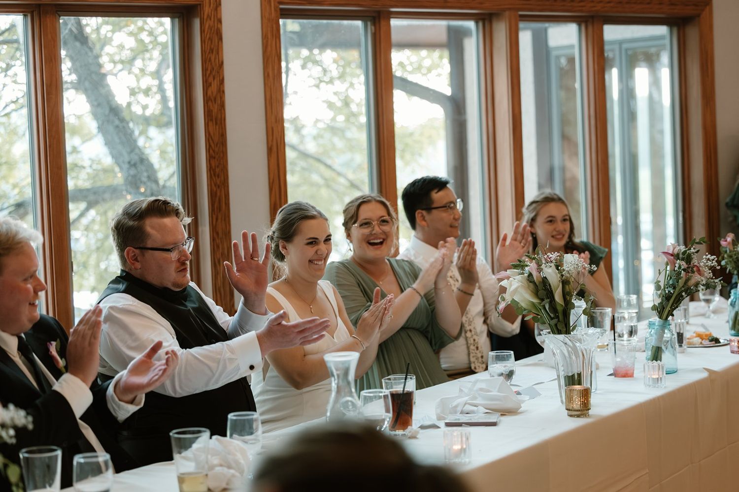 Wedding guests seated at a long table applaud during a reception celebration in a bright windowed room.