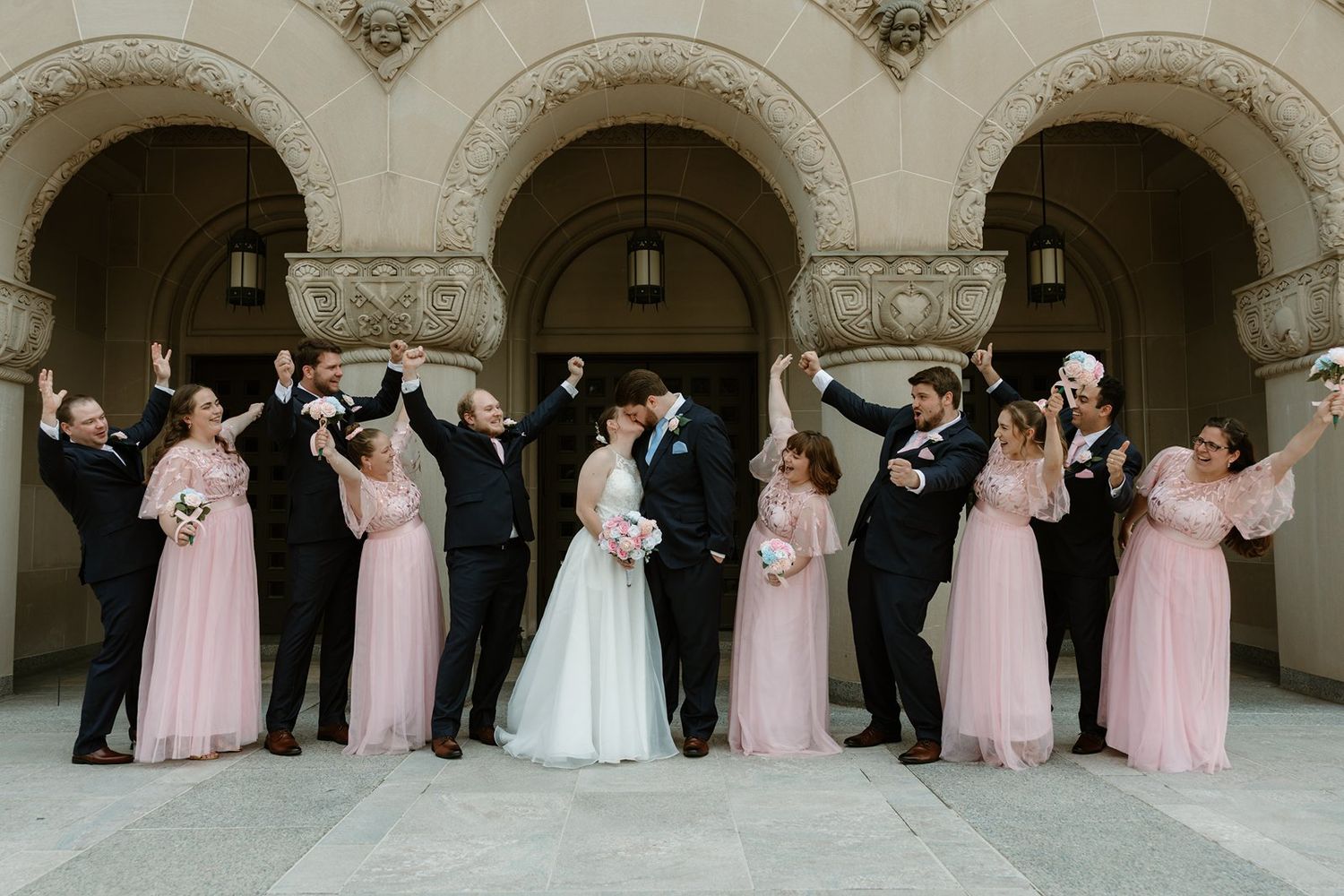 Wedding party poses under ornate stone archways wearing pink bridesmaid dresses and formal attire.