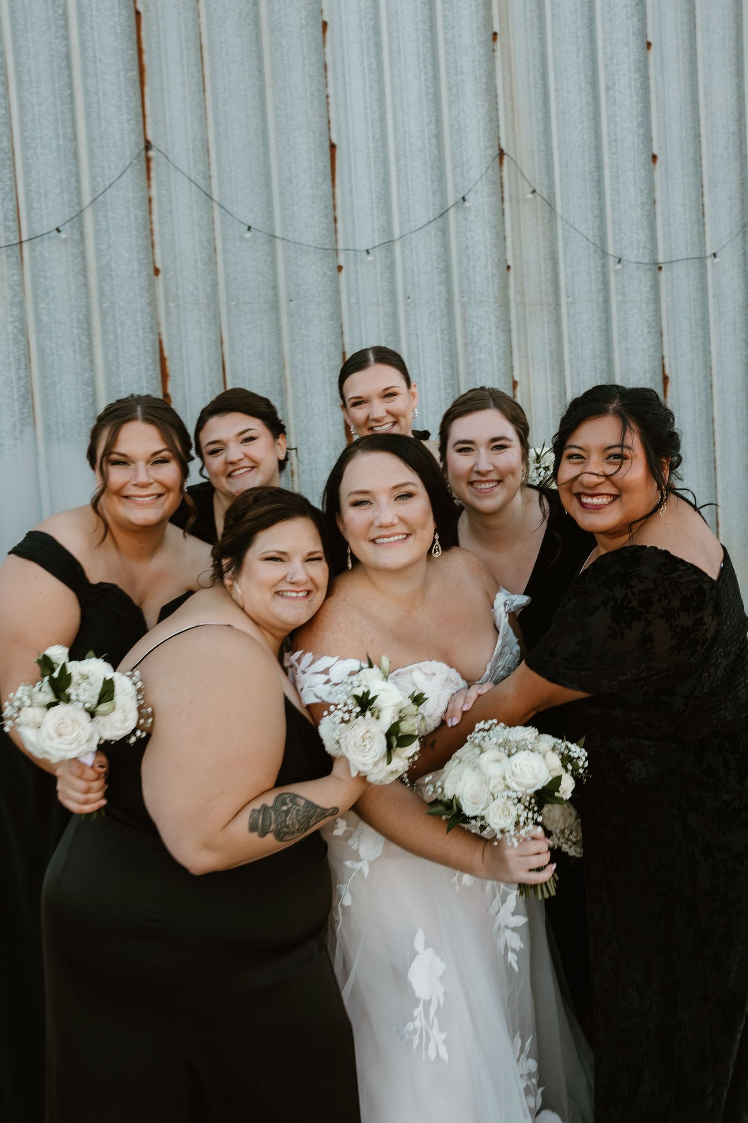 A bridal party in black dresses and a white wedding gown pose together holding white floral bouquets against a metal wall.