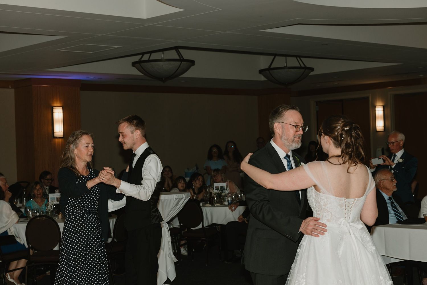 Couples dancing together at an indoor wedding reception with tables and decor visible in the background.