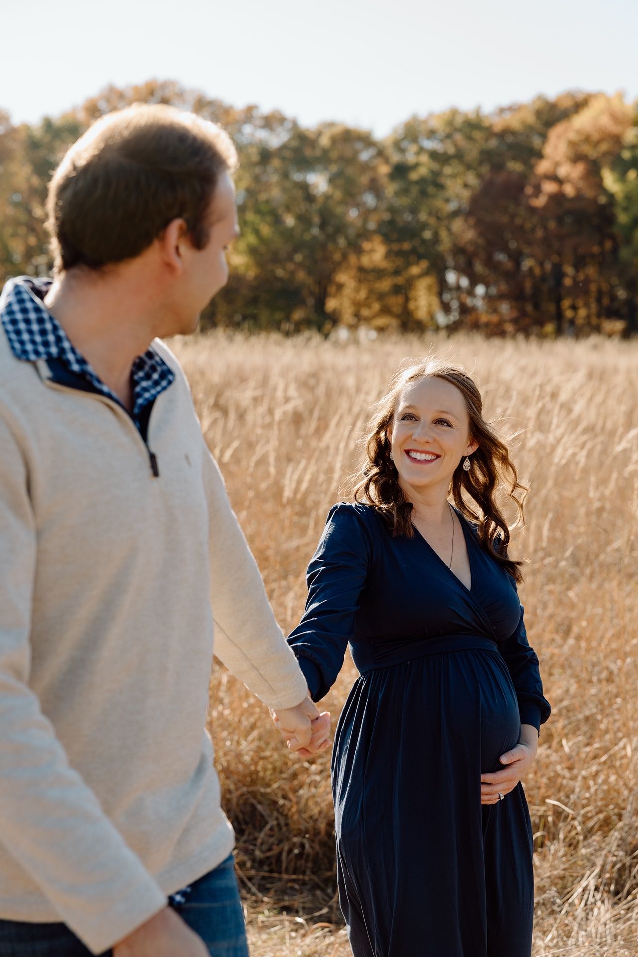 Pregnant woman in navy dress holds hands with man in field during autumn maternity photoshoot.