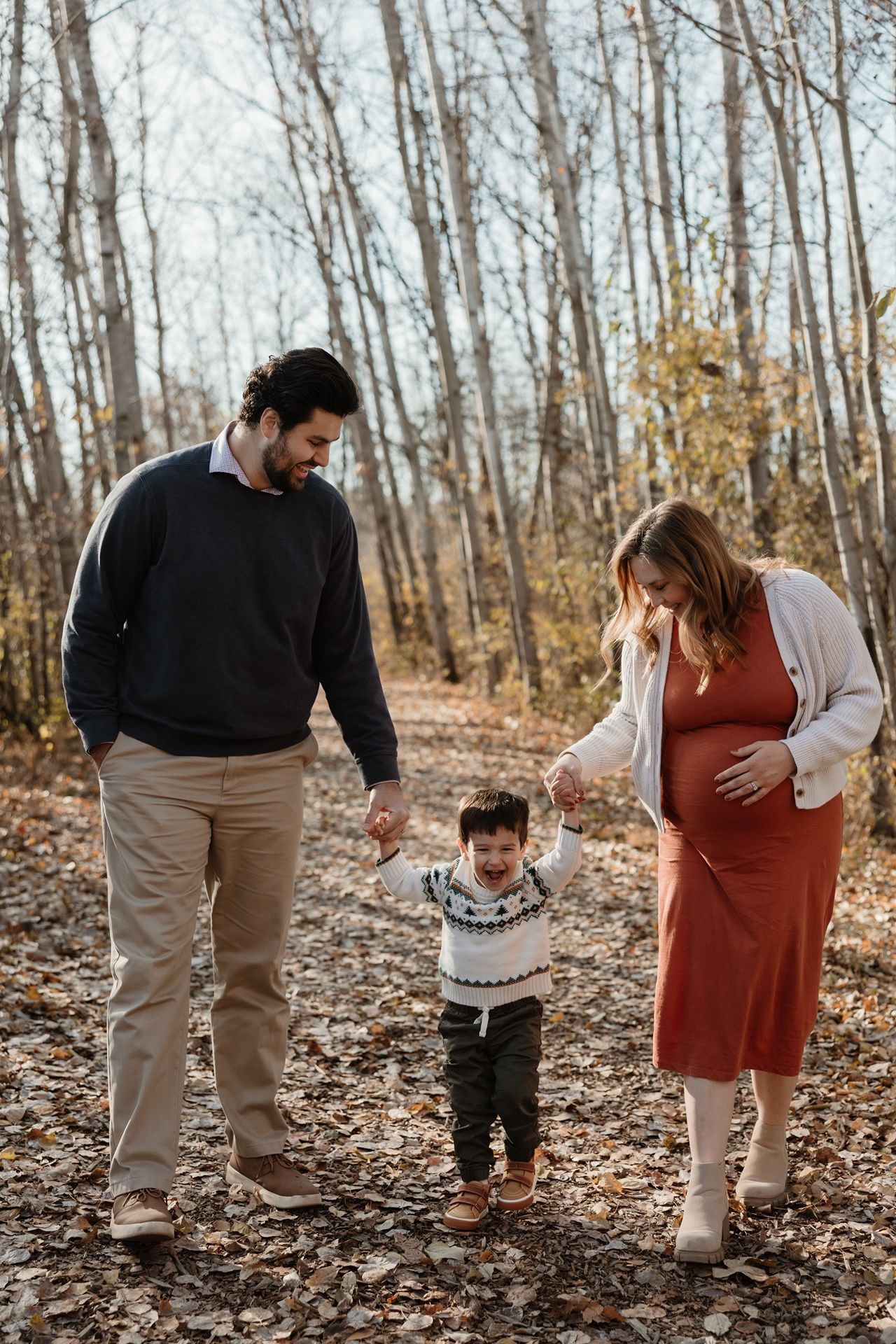 Pregnant family walking in autumn forest, parents holding toddler's hands among fall leaves.