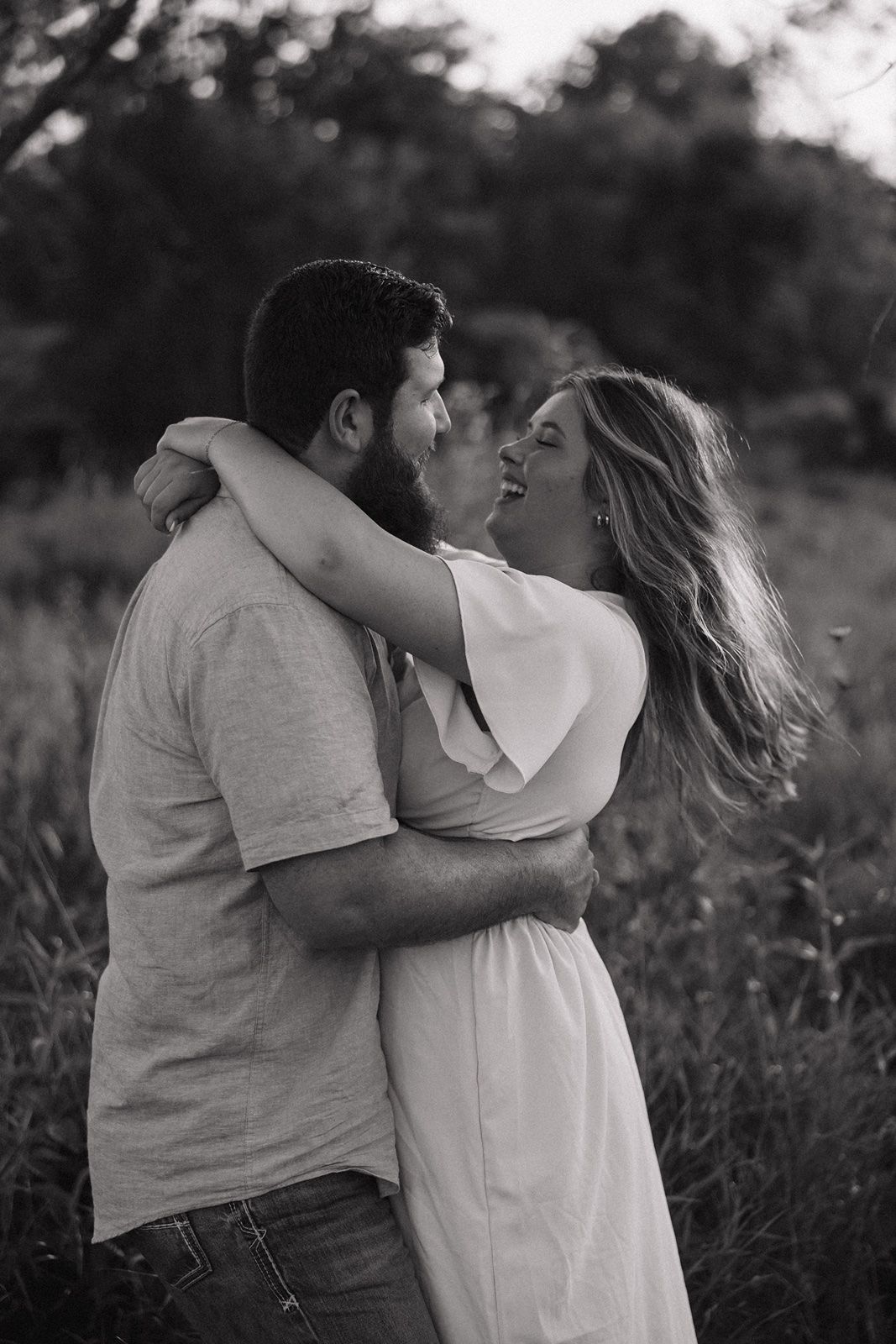 Black and white photo of a couple embracing and laughing together in a field, woman in white dress with flowing hair.