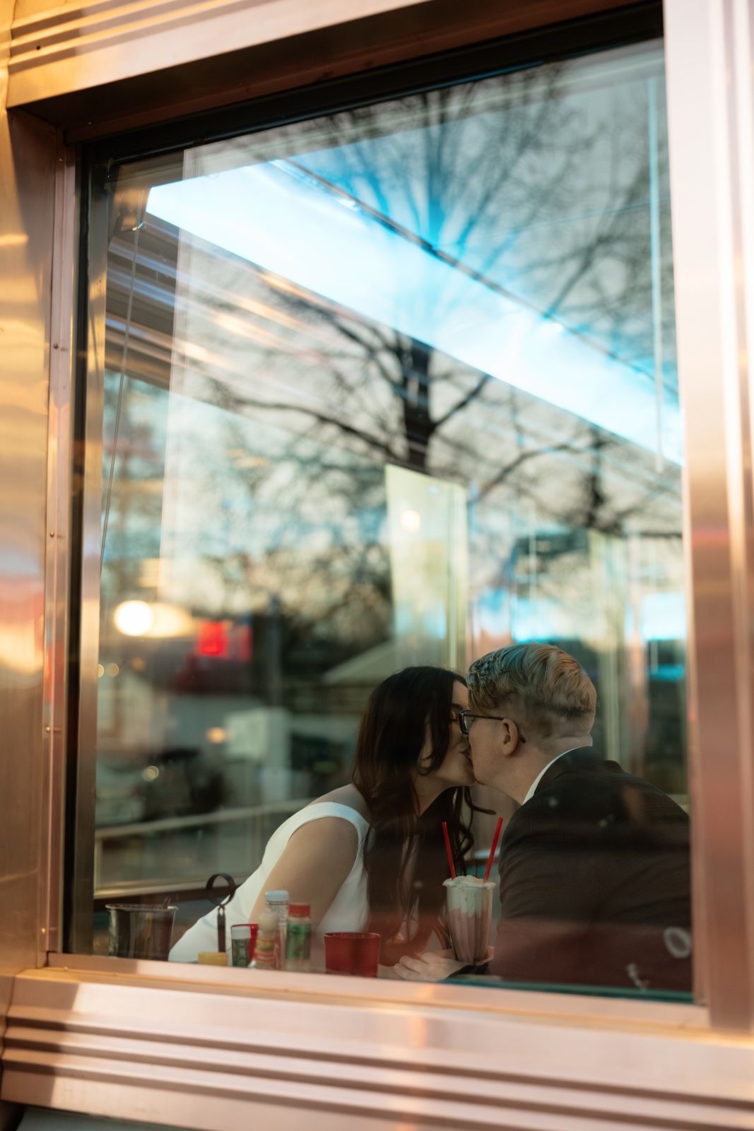 Couple sharing a romantic kiss inside a diner booth, viewed through a window with tree reflections.