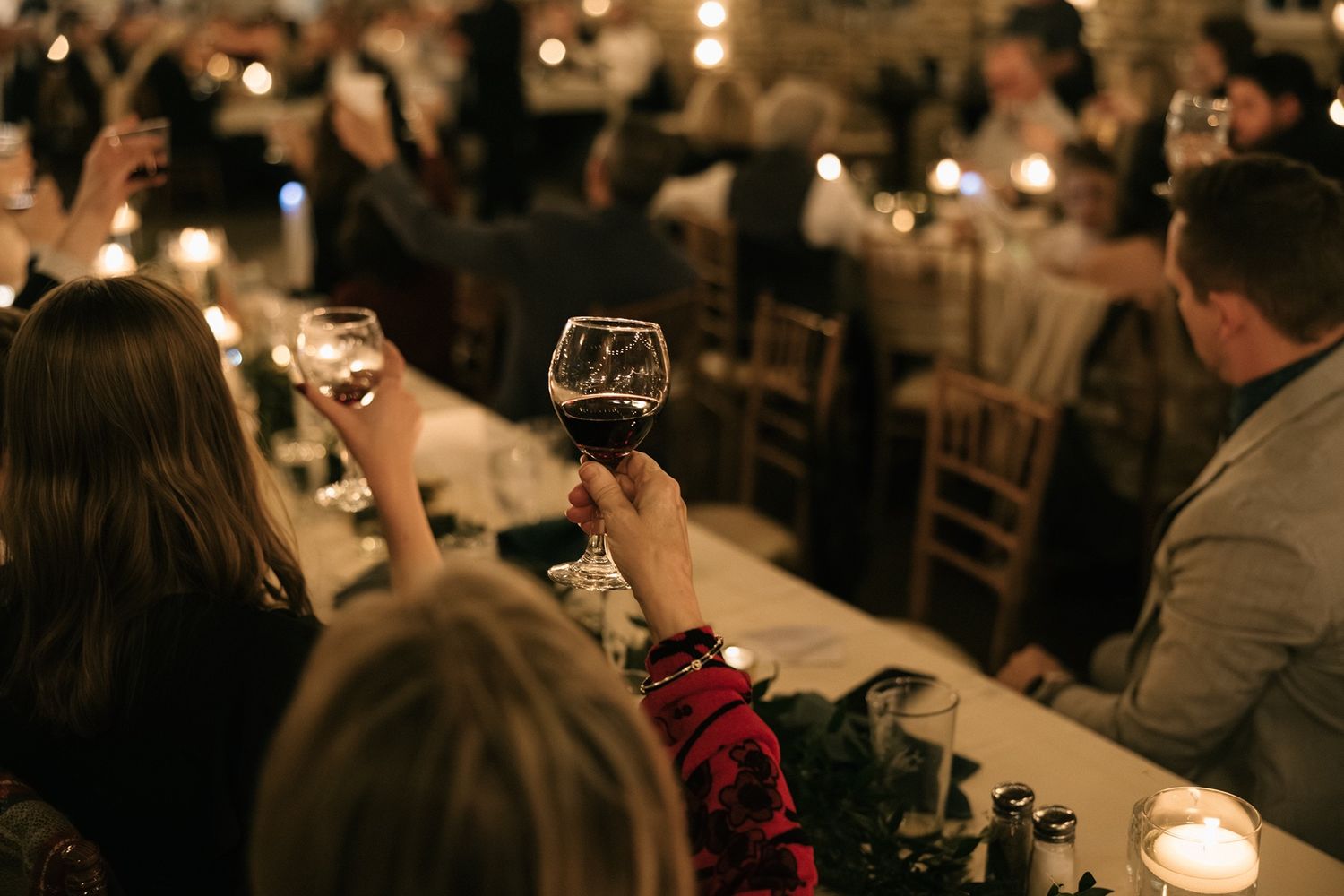 Guests raising wine glasses in a candlelit dinner reception hall during a festive evening celebration.