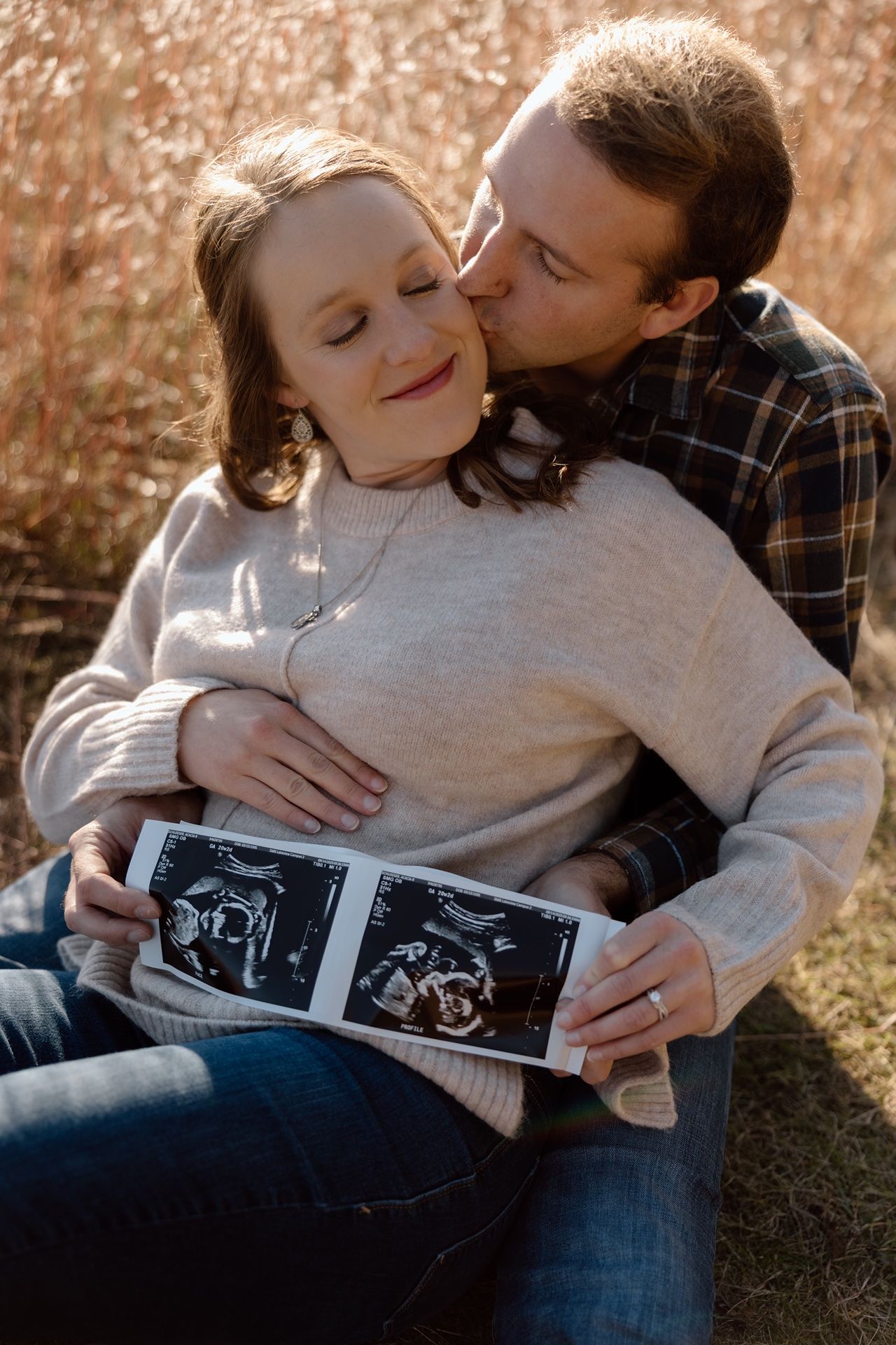 Couple sitting outdoors holding ultrasound photos, man kissing woman on cheek, announcing pregnancy in golden field.