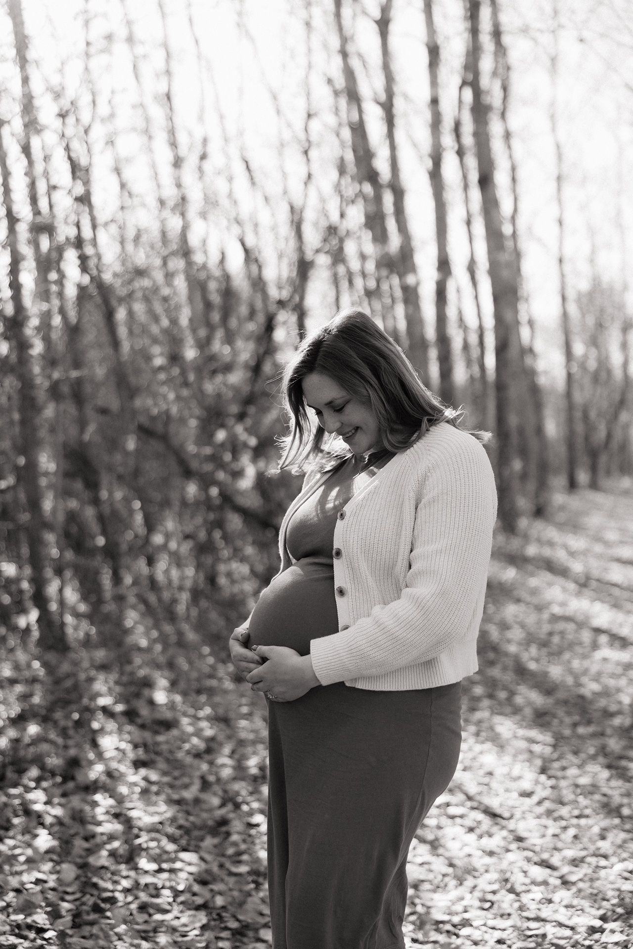 Black and white photo of a pregnant woman cradling her baby bump in an autumn forest setting.