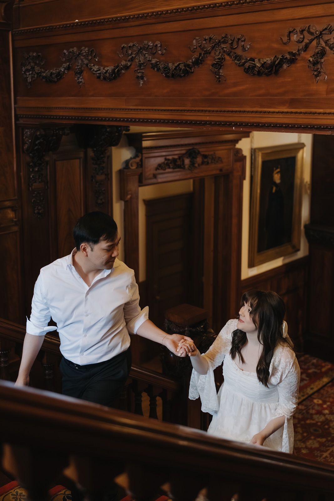 Couple holding hands on ornate wooden staircase in historic mansion, man in white shirt, woman in white dress.