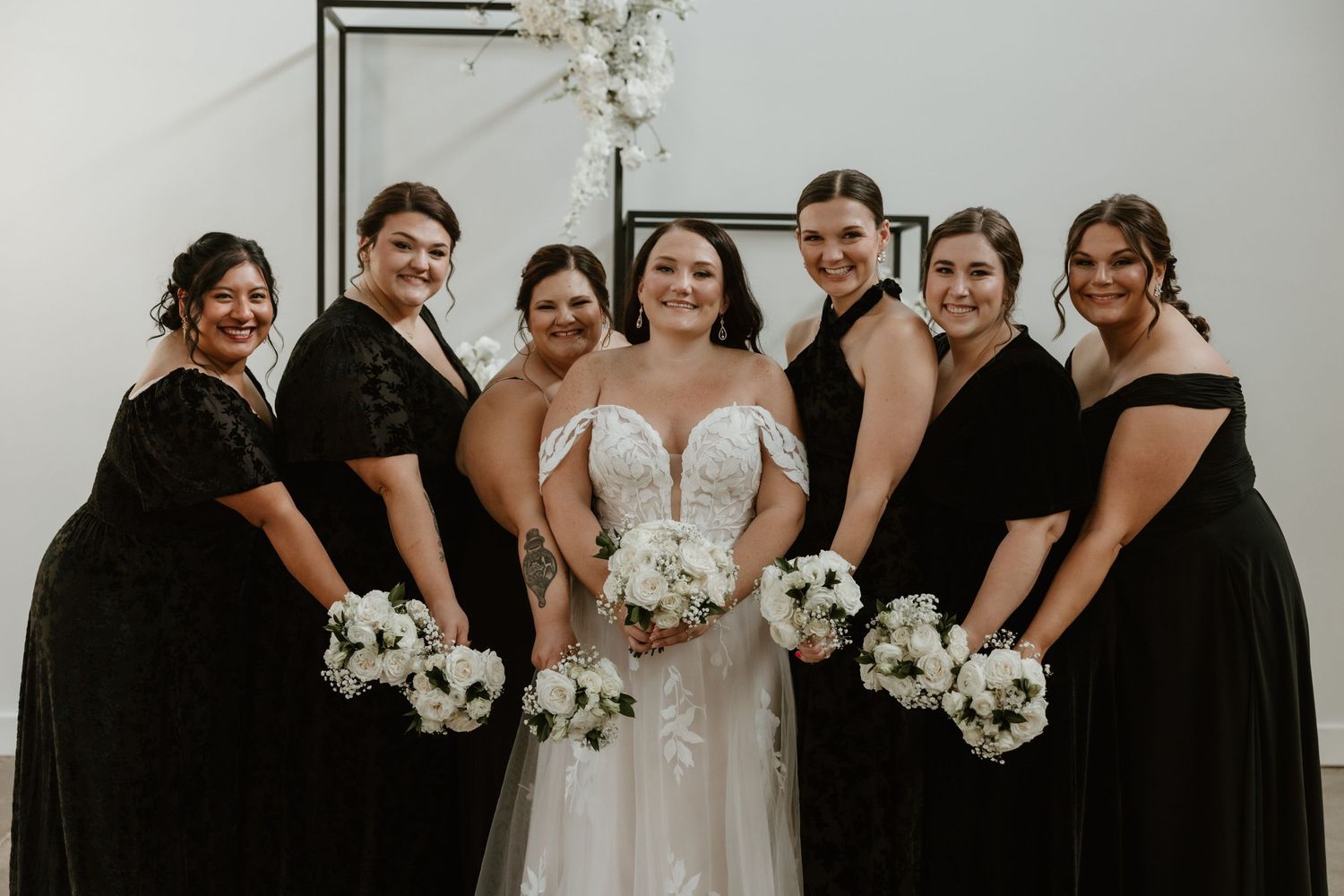 A group in black dresses stands with a person in a white wedding gown holding bouquets against a light background.