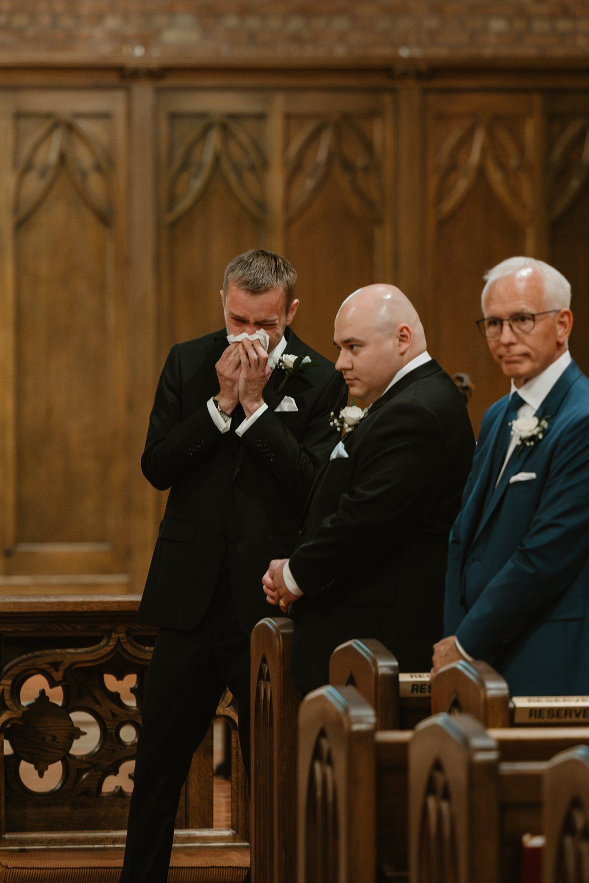 Three men in formal attire stand at the altar during an emotional moment in a church with ornate wooden panel walls.